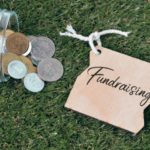 A glass jar tipped over, spilling coins on grass next to a wooden tag labeled 'Fundraising'.